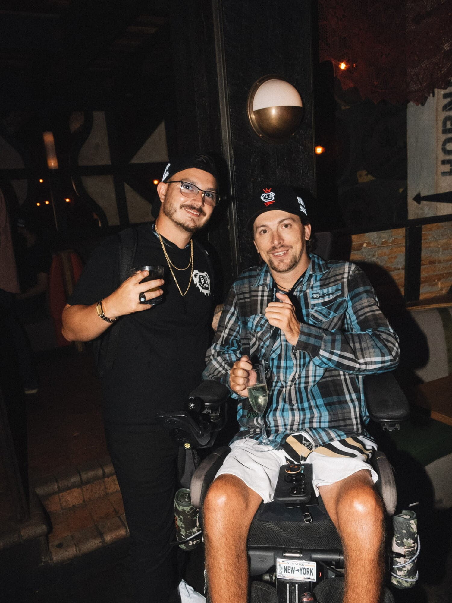 Jag (JagOBX) in a black cap and tee, holding a drink, standing with a friend outside a venue at night