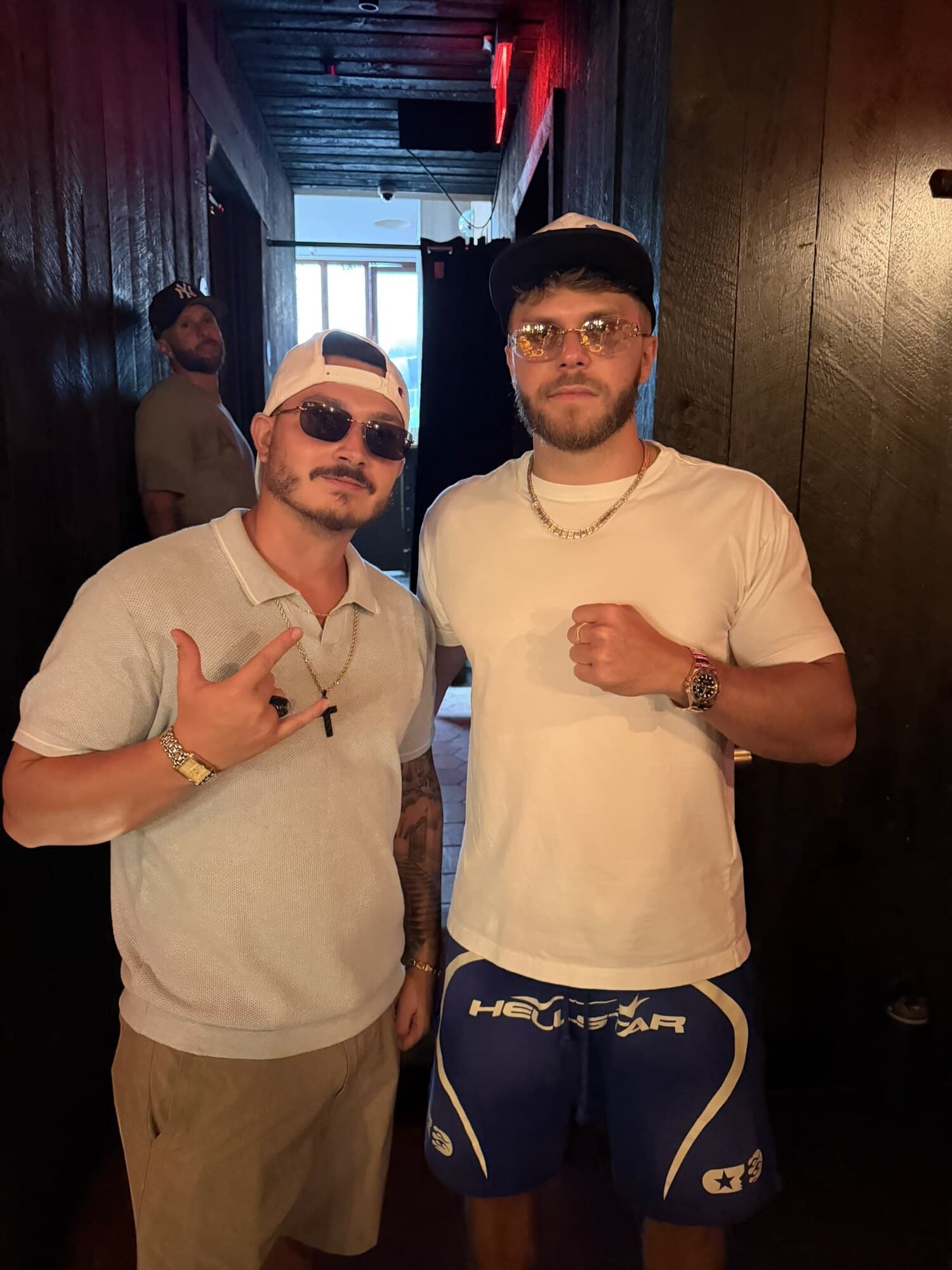 Jag (JagOBX) in a cream polo, backwards cap, and sunglasses standing with a friend inside a venue